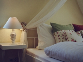 Das romantische Schlafzimmer mit einem weißen Metallbett und eingefasstem Betthimmel.
Ebenso hochwertigen Stores und Seitenschals mit Rosendekor.